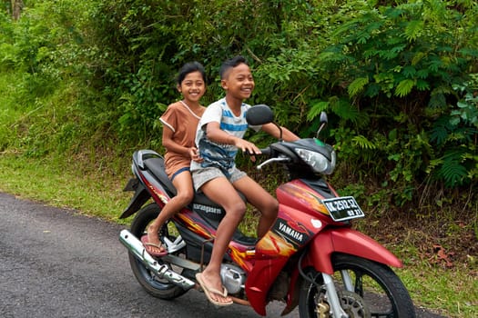 Children ride a scooter without helmets on a road in a village in Asia. Bali, Indonesia - 12.24.2022