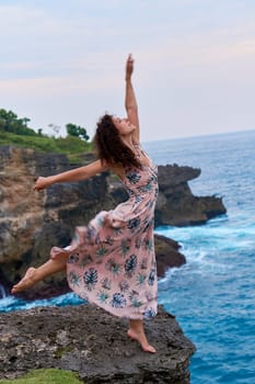 A pretty slender woman in a long dress poses on a rock on a cliff above the ocean