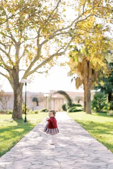 Little girl stands half-turned on a paved path in the park and waves her hand affably. High quality photo