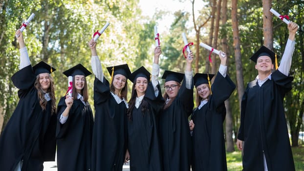 Row of young students in graduation gowns outdoors showing off their diplomas