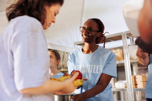 Portrait of female charity worker handing out fresh fruits and food to poor and homeless people. Cheerful black woman volunteers at food drive serving free meals to the hungry and less fortunate.