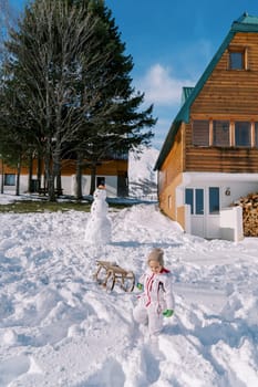 Little girl drags a sled on a rope from a hill near a wooden chalet. High quality photo