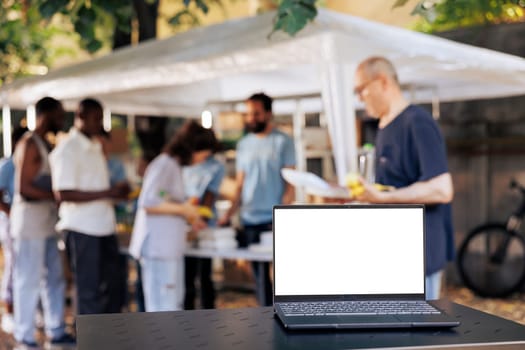 Picture shows digital laptop with a blank white screen that is sitting on a table while charity workers help the less fortunate. Minicomputer in the foreground with isolated copyspace mockup template.