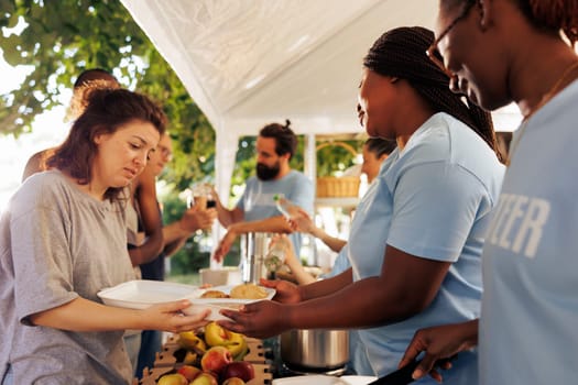 Hunger relief team of volunteers donates essential items and free food to needy, showing generosity in serving less fortunate. Non-profit outdoor food bank sharing warm meals to poor homeless people.