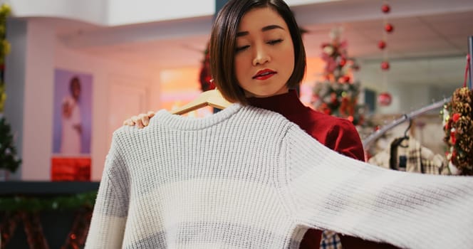 Dolly in shot of asian customer in xmas decorated clothing store looking to buy comfortable garment on clearance, taking advantage of Christmas promotional sales during winter holiday season