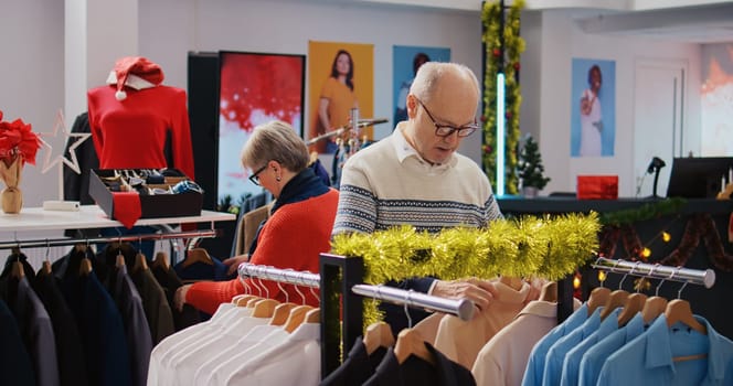 Senescent customers browsing through clothing racks in festive ornate fashion boutique during xmas holiday season. Aged couple happy after finding colorful blazers to gift at Christmas family event