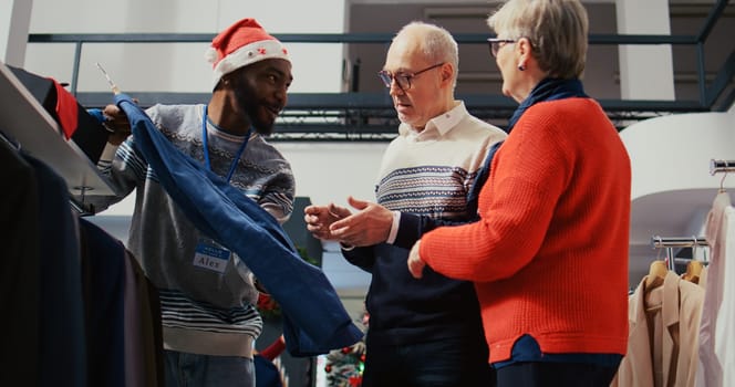 Elderly couple in festive adorn shopping mall fashion boutique, being helped by friendly retail assistant with finding ideal outfit during Chrismas holiday season promotional sales