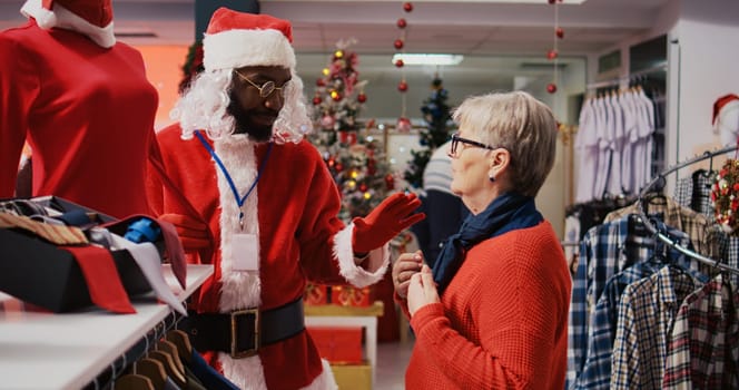 Employee wearing Santa Claus costume in busy shopping mall fashion boutique chatting with older woman. Worker in holiday themed suit showing aged customer red clothing piece