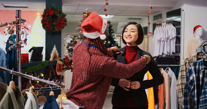 Asian client getting out of changing room, excited about elegant blazer fitting perfectly, speaking with helpful retail assistant in Christmas themed clothing store during winter holiday season