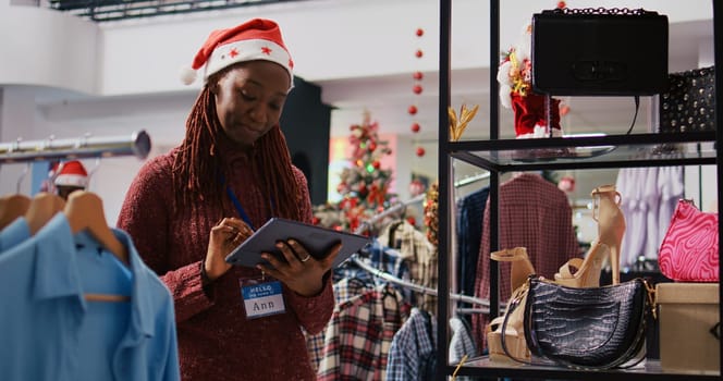 African american supervisor wearing Santa hat browsing through clothes racks at Christmas themed fashion boutique, using tablet to input promotional prices on online store during holiday season sales