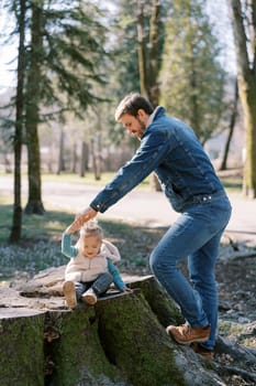 Dad stands near a huge stump in the forest with a little girl sitting on it holding her hand. High quality photo