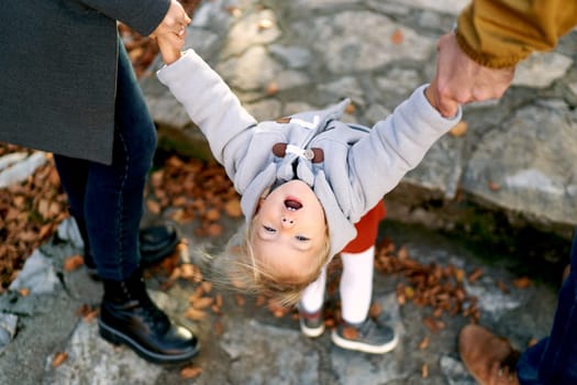 Little girl stands with her head thrown back holding her parents hands on a paved path in the park. Top view. Cropped. High quality photo