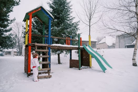 Little girl stands near a snow-covered colorful slide among the trees and looks to the side. High quality photo