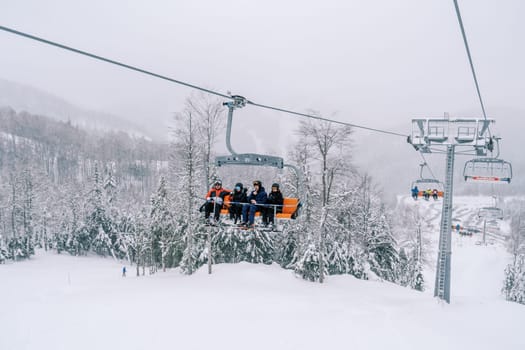 Skiers in bright suits ride on a chairlift over a snowy forest up in the mountains. High quality photo