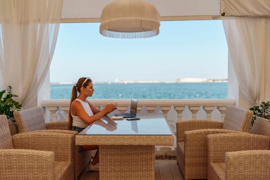 Woman coffee cafe laptop sea. Female freelancer using laptop on coffee break in modern cafe with sea view in the morning.