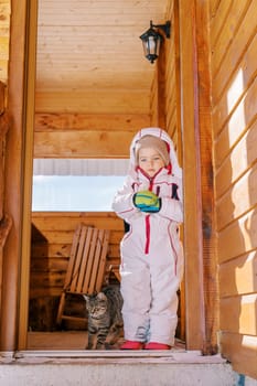 Little girl stands with a tabby cat on the threshold of a wooden cottage and looks down. High quality photo
