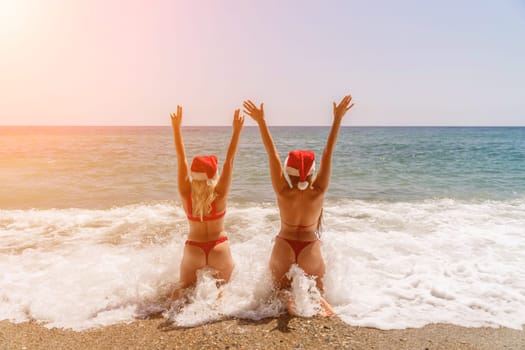 Women Santa hats ocean play. Seaside, beach daytime, enjoying beach fun. Two women in red swimsuits and Santa hats are enjoying themselves in the ocean, kneeling in the waves and raising their hands up