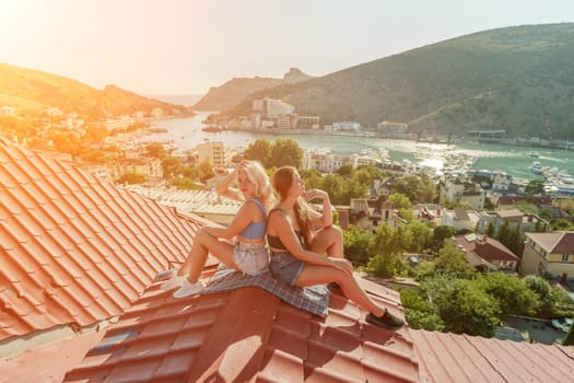 Two women sitting on a red roof, enjoying the view of the town and the sea. Rooftop vantage point. In the background, there are several boats visible on the water, adding to the picturesque scene