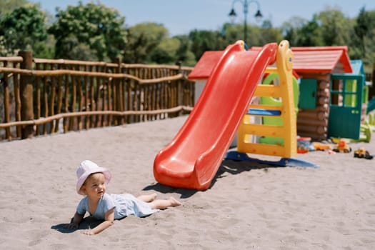 Little girl lies on the sand near a slide on the playground and looks away. High quality photo