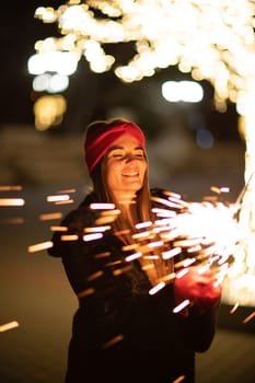 Woman holding sparkler night while celebrating Christmas outside. Dressed in a fur coat and a red headband. Blurred christmas decorations in the background. Selective focus.