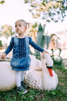 Little girl stands near a sculpture of a swan, putting her hand on his neck. High quality photo