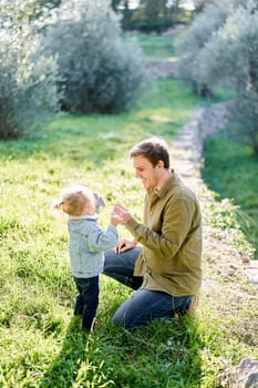 Dad is squatting near a little girl with a dandelion in his hand in a clearing. High quality photo