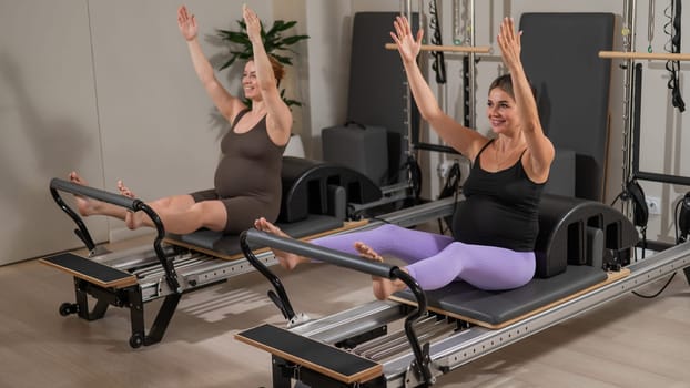 Two pregnant women do Pilates exercises on a reformer. Yoga class for pregnant women