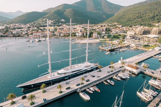Long sailing yacht is moored at the pier against the background of the coast with modern hotels and high-rise buildings. Porto, Montenegro. Drone. High quality photo