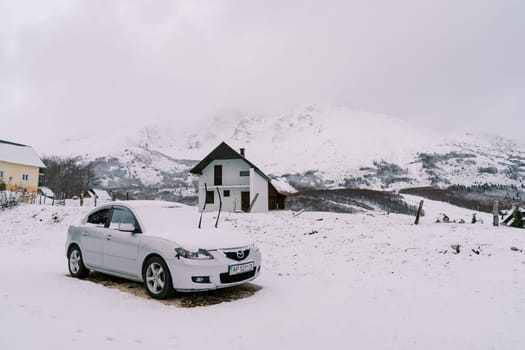 Snow-covered car with raised windshield wipers stands near a cottage in a mountain valley. High quality photo