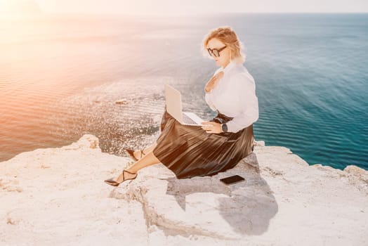 Digital nomad, Business woman working on laptop by the sea. Pretty lady typing on computer by the sea at sunset, makes a business transaction online from a distance. Freelance, remote work on vacation