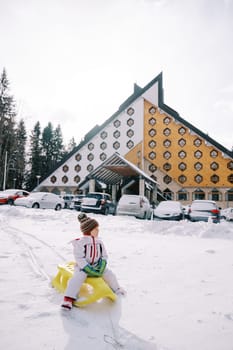 Little girl sits on a sled in the snow near cars parked near a triangular hotel. High quality photo