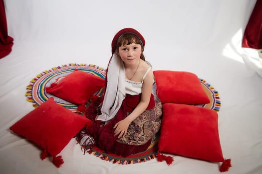 Portrait of Little girl in a stylized Tatar national costume having rest with a red pillow on a white background in the studio. Photo shoot of funny young teenager who is not a professional model