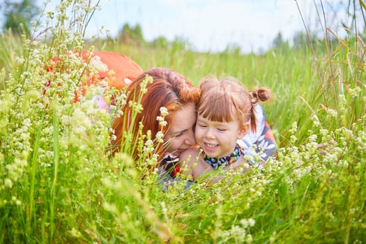 Happy female family with mother and daughter on green and yellow meadow full of grass and flower. Woman with red hair and blonde girl having fun, joy and hug in sunny summer day. Concept family love