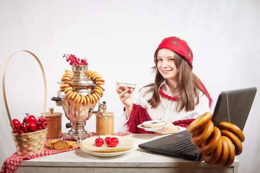 Girl in a red colored scarf, white shirt and shawl near a table with a samovar, bagels drinks tea for the Easter holiday and Masolennitsa. A freelancer or blogger with a laptop is resting and working