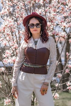 Magnolia park woman. Stylish woman in a hat stands near the magnolia bush in the park. Dressed in white corset pants and posing for the camera