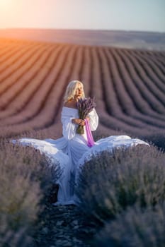 Blonde woman poses in lavender field at sunset. Happy woman in white dress holds lavender bouquet. Aromatherapy concept, lavender oil, photo session in lavender.