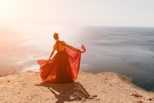 Side view a Young beautiful sensual woman in a red long dress posing on a rock high above the sea during sunrise. Girl on the nature on blue sky background. Fashion photo.