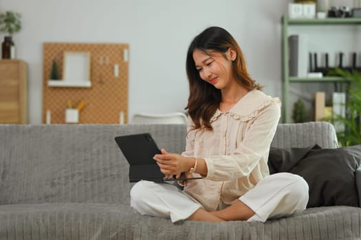 Happy young woman dressed in casual clothes sitting on couch and browsing internet on digital tablet