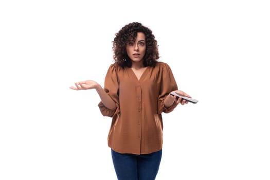 young upset slender woman with curls dressed in a brown shirt shrugs her shoulders holding a phone in her hand.
