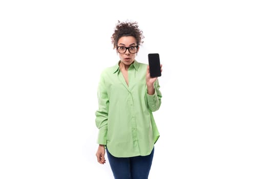 young pretty european brunette curly woman dressed in a light green shirt and glasses holds a smartphone.
