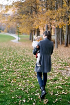 Mom with a little girl in her arms walks along the green grass in the park. Back view. High quality photo
