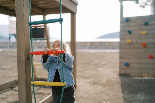 Little girl stands near a rope ladder on a playground and looks up. High quality photo