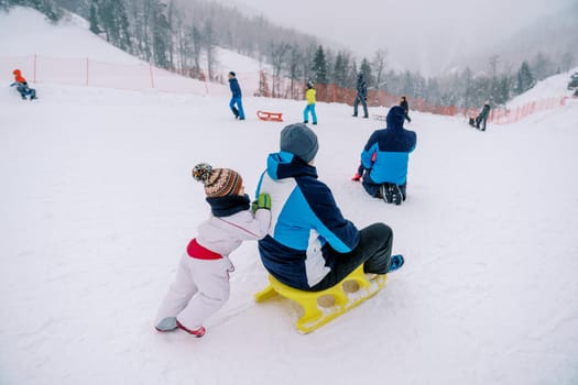 Small child pushes his dad on a sled down a snowy hill. Back view. High quality photo