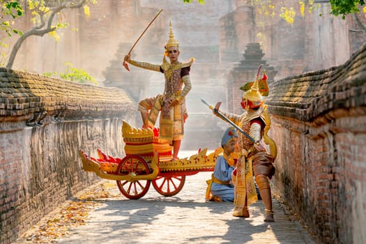 Asian man with old Thai traditional cloth hold weapon and stand and action of dance on traditional chariot also hold weapon action to fight with red giant in front of ancient wall and building.