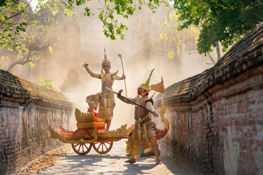 Asian man with old Thai traditional cloth hold weapon and stand and action of dance on traditional chariot also hold weapon action to fight with red giant in front of ancient wall and building.