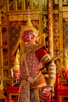 Close up Khon or traditional Thai classic masked from the Ramakien with Red giant character stand with spear and look forward with Thai painting wall in background.