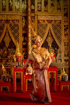 Vertical image of Asian woman wear Thai ancient traditional dress action of dancing in front of Khon or traditional Thai classic masked from the Ramakien on wooden shelves and painting.