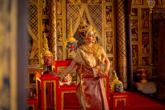 Side view of beautiful Asian woman wear Thai ancient traditional dress action of dancing in front of Khon or traditional Thai classic masked from the Ramakien and painting on public wall.