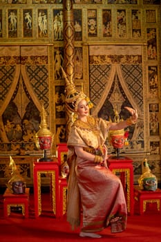 Beautiful Asian woman wear Thai ancient traditional dress action of dancing in front of shelves with Khon masks and Thai painting as background in public place.