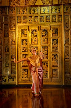 Vertical image of beautiful Asian woman wear Thai ancient traditional dress action of dancing in front of Thai painting as background in public place.